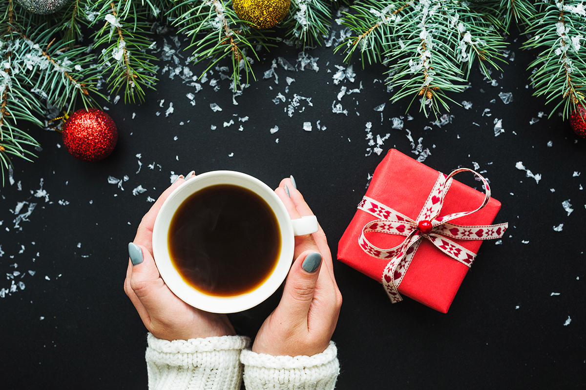 Image of hands holding coffee by a present and a christmas tree